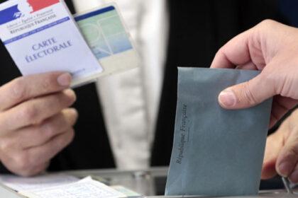 L'enveloppe électorale est mise dans l'urne. En arrière-plan, un homme présente sa carte électorale et sa pièce d'identité