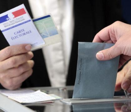 L'enveloppe électorale est mise dans l'urne. En arrière-plan, un homme présente sa carte électorale et sa pièce d'identité
